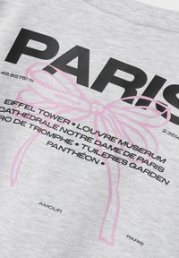 Felpa grigia con grande testo nero "PARIS", grafica di un fiocco rosa, e nomi di luoghi con coordinate e "AMOUR" in basso.