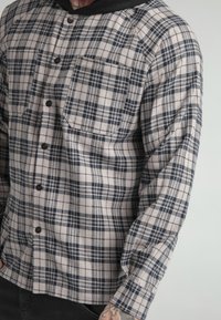 Camicia a quadri grigia e blu navy, con due tasche sul petto, un cappuccio nero e una vestibilità comoda con maniche raglan.