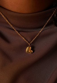 Gouden hartvormige hanger aan een delicate ketting, die rust op een donkerbruine textuurstof. De hanger heeft een glad, reflecterend oppervlak.
