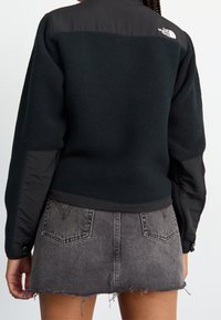 Svart fleecetröja med ett slätt svart tygöverdrag på axlarna. Har en kort design och synliga sömnadsdetaljer.