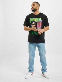 Camiseta de algodón negra con un estampado gráfico colorido que presenta un rostro y texto. Combinada con jeans azul claro y zapatillas blancas con acentos rojos.