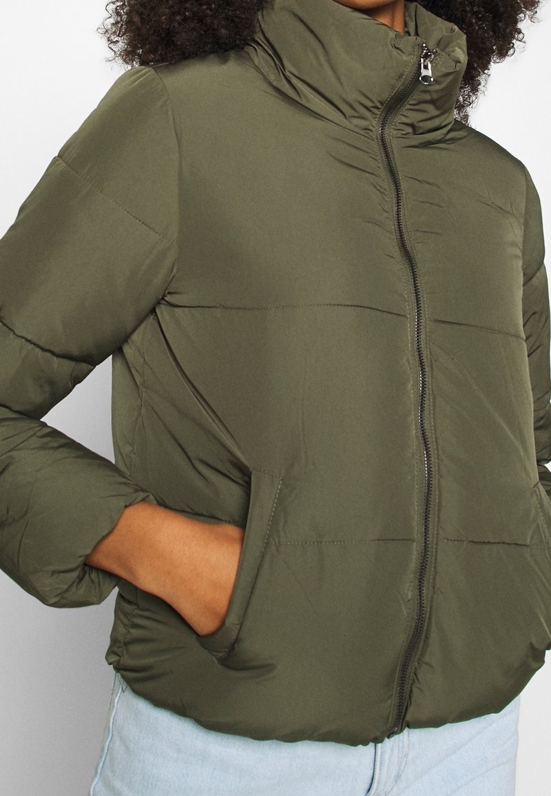 Personne portant une doudoune vert olive avec un col montant et une fermeture éclair devant, mains dans les poches de la veste, associée à un jean bleu clair.