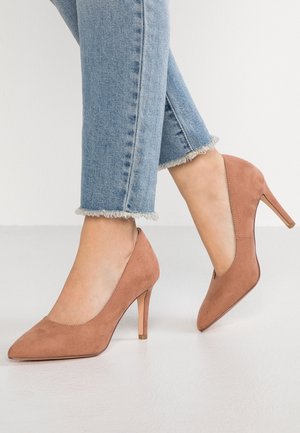 Person iført lyseblå cropped jeans og brune ruskindspointede højhælede sko, stående på et hvidt gulv.