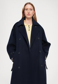 Meripihka wool trench coat dengan desain ganda, kerah besar, dan tombol hitam; tekstur lembut; dikenakan di atas kaos polo kuning.
