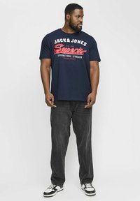 Camiseta de algodón azul marino con texto blanco y rojo. Combinada con jeans gris oscuro y zapatillas de deporte en blanco y negro. Ajuste estándar con mangas cortas.