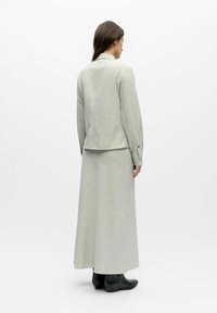 Femme de dos portant une veste gris clair à manches longues et une jupe longue assortie, avec des bottines noires, sur fond blanc.