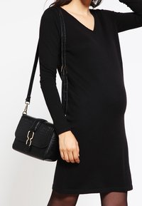 Robe noire à manches longues avec un décolleté en V, silhouette ajustée. Tient un sac à main noir texturé avec des accessoires en doré et une bandoulière.