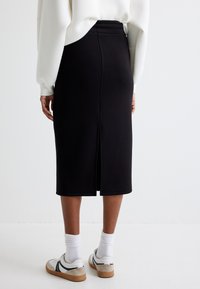 Zwarte midi-jurk gemaakt van elastische stof, met een split aan de achterkant en een hoge tailleband, gecombineerd met witte sneakers en geribbelde enkelsokken.