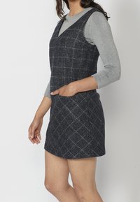 Navy plaid sleeveless dress made of textured fabric, featuring a V-neck and side pockets, worn over a light gray long-sleeve top.