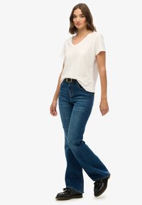 Camiseta de algodón blanca con cuello en V combinada con jeans acampanados azules, que llevan un cinturón negro y zapatos negros, todo sobre un fondo blanco liso.