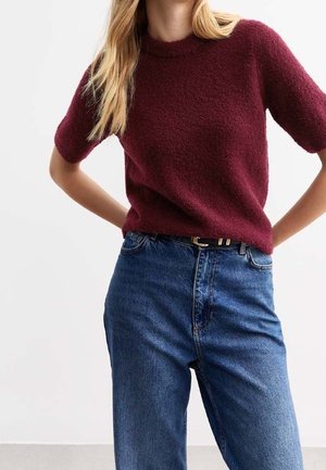 Vrouw draagt een bordeauxrode kortemouwen gebreide trui, ingestopt in een high-waisted blauwe spijkerbroek met een zwarte riem, staand met haar handen op haar heupen.