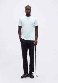 Άνδρας που στέκεται κρατώντας ένα μπαστούνι του γκολφ, φορώντας ένα ανοιχτό μπλε T-shirt Calvin Klein, μαύρο παντελόνι και μαύρα αθλητικά παπούτσια σε απλό λευκό φόντο.