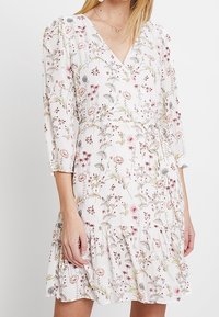 Floral wrap dress with a white base, featuring pink and green flower patterns, three-quarter sleeves, a cinched waist, and a ruffled hem.