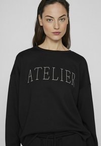 Juodas sportinis megztinis su užrašytu "ATELIER" baltomis raidėmis, laisvas kirpimas, apvalus iškirpimas ir minkšta tekstūra. Ilgos rankovės ir paprastas dizainas.