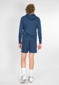 Felpa navy con tasca a marsupio e lacci, abbinata a pantaloni corti navy con un lato a fantasia colorata. Calze e scarpe da ginnastica bianche.