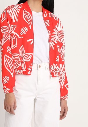 Frau trägt eine rote Reißverschlussjacke mit weißen Blumenmustern über einem weißen Hemd und weißen, hoch taillierten Hosen.