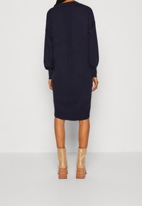 Robe bleu marine à manches longues avec des détails côtelés aux épaules et dans le dos, associée à des bottines beiges texturées à talons carrés.