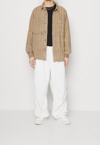 Rutigt beige och brunt skjorta med knappar över en svart t-shirt, kombinerad med vita baggy byxor och vita sneakers. Subtila textur- och designdetaljer.