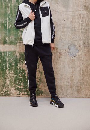 Personne portant un survêtement noir Nike avec un gilet en polaire blanc et des baskets noires, debout contre un mur texturé vert et beige usé.