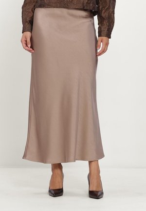 Vrouw draagt een gladde beige midi rok gecombineerd met donkerbruine puntige hoge hakken en een bruin gestructureerde top met lange mouwen.