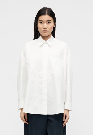 Kobieta z długimi czarnymi włosami ubrana w oversize'ową białą koszulę na guziki i ciemne spodnie, stojąca na tle gładkiego, jasnoszarego tła.