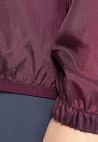 Chaqueta ligera burdeos con tejido texturizado, puños y cinturilla elásticos. Detalles de costura visibles a lo largo del dobladillo.