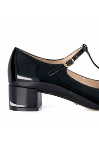 Chaussure à talon bloc en cuir verni noir avec un bout arrondi, dotée d'une fine sangle à la cheville et d'une accent en boucle dorée.