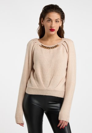Vrouw met donker haar draagt een beige trui met gouden kettingdetail en zwarte leren broek, staand tegen een effen achtergrond.