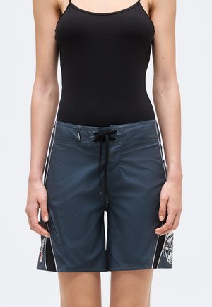 Kvinde iført mørkeblå knælange badeshorts med sort snor og sidestriber, kombineret med en sort ærmeløs top.