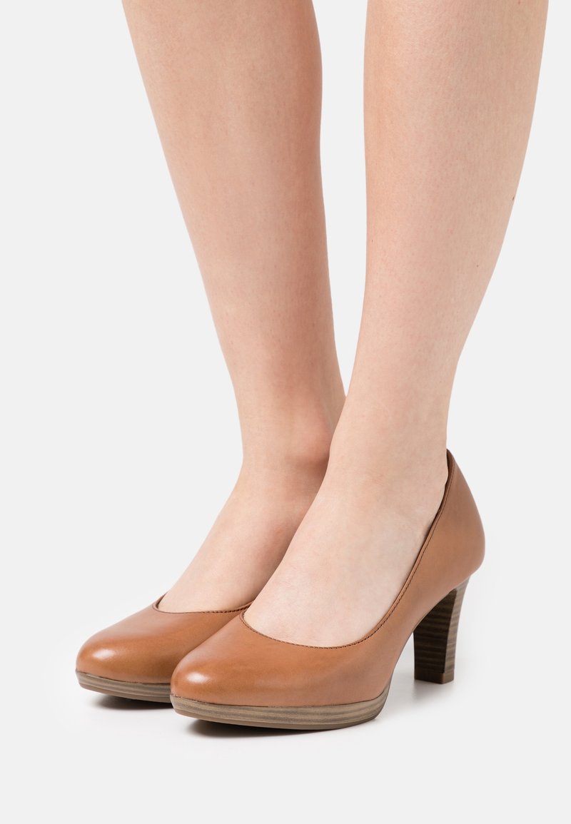 Jambes portant des chaussures à talons hauts en cuir marron clair avec des bouts arrondis et des talons empilés sur un fond clair uni.