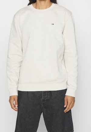 Person trägt ein cremefarbenes Sweatshirt mit Rundhalsausschnitt und kleinem Logo auf der linken Brust sowie schwarze Jeans und steht vor einem schlichten Hintergrund.