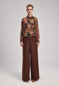 Bluse med blomsterprint, halvt gennemsigtig i jordfarver, parret med brede brune bukser. Bemærkelsesværdige detaljer inkluderer knaplommer og et bånd i taljen.