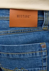 Jeans de mezclilla azul oscuro con una prominente etiqueta de cuero marrón marcada "MUSTANG" y detalles de costura amarilla a lo largo de las costuras de los bolsillos traseros.