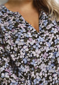 Frau in einer V-Ausschnittbluse mit dichtem Blumenmuster in Blau-, Lila-, Weiß- und Grüntönen auf schwarzem Hintergrund.