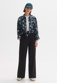 Pantaloni neri a gamba larga abbinati a una camicia blu a fantasia con forme tonde e frontale annodato, completati da un cappello verde e scarpe.
