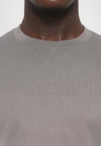 Pelēka ribota T-kreklā ar izspiestu tekstu "ARMANI EXCHANGE MILANO/NEW YORK" uz krūtīm. Apaļš kakls, pieguļoša forma.