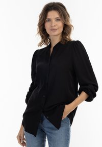 Zwarte blouse met knopen en een zachte textuur, geplooide mouwen en een kraag. Gepaard met lichtblauwe jeans, die een ontspannen pasvorm laten zien.
