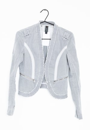 Veste blazer pour femmes gris clair avec surpiqûres blanches et poches zippées, suspendue sur un cintre noir contre un fond blanc.