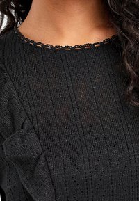 Pull en tricot noir avec une texture côtelée. Présente un décolleté en scallop et un détail volanté à l'épaule. Design léger et ajusté.