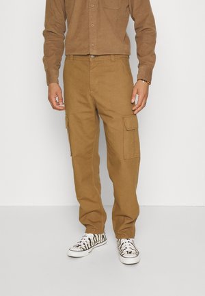 Man draagt lichtbruine cargobroek met zijzakken, een bijpassend lichtbruin overhemd met knopen en witte sneakers met zwarte zebrastripes.