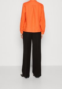 Chemise orange à manches longues en lin avec un détail dans le dos froncé, assortie à un pantalon large noir taille haute et des talons noirs.