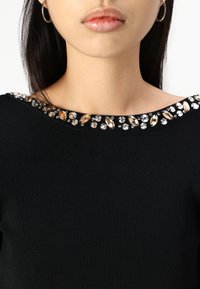 Svart ribbestrikket topp med vid halsringning som har en dekorativ kant av klare og ravfargede krystaller langs kragen.