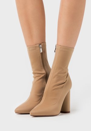 Bottes beiges montant à la cheville avec des bouts pointus et des talons carrés hauts, portées sur des jambes nues, dotées de fermetures éclair latérales et d'une texture de tissu lisse.
