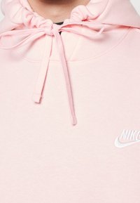 Ljusrosa hoodie gjord av mjukt material, med en snörning vid halsringningen och en vit broderad Nike-logotyp.