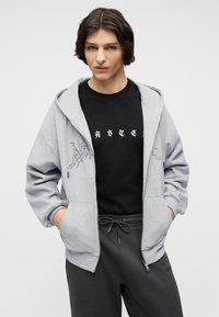 Νεαρό άτομο που φοράει γκρι φούτερ με φερμουάρ, μαύρο μπλουζάκι με σχέδιο και σκούρο γκρι παντελόνι με κορδόνι, στέκεται με τα χέρια στις τσέπες.