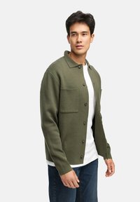 Grön strukturerad cardigan med klassisk krage, framknappar och två bröstfickor, lagd över en vit t-shirt, i kombination med blå jeans.