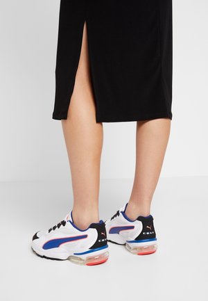 Osoba ubrana w czarną spódnicę z bocznym rozcięciem i białe sneakersy Puma z niebieskimi i czerwonymi akcentami, stojąca na jasnoszarej podłodze.