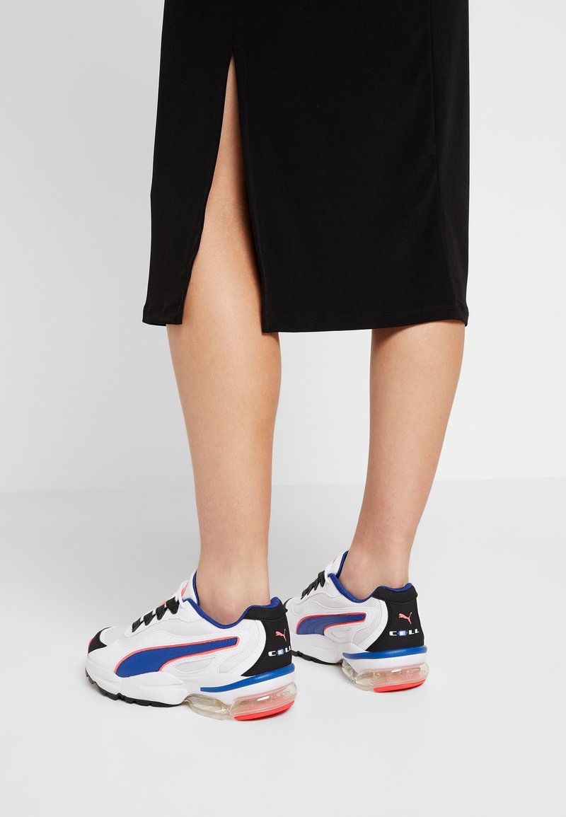 Personne portant une jupe noire avec une fente latérale et des baskets blanches Puma avec des touches bleues et rouges, debout sur un sol gris clair.