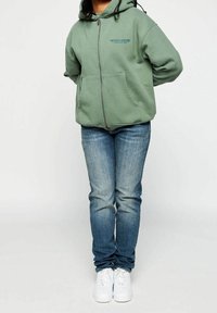 Veste à capuche zippée verte avec de grandes poches avant, en mélange de coton, portée sur un jean en denim bleu délavé. Des baskets blanches complètent le look.