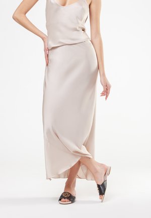 Donna che indossa un vestito sottoveste in raso beige chiaro con sandali neri bassi con punta aperta e un emblema dorato, in piedi con una gamba incrociata sull'altra.